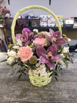 Плетеная корзинка с фиолетовыми орхидеями, гвоздиками, альстромерями, хризантемами и декоративной зеленью
